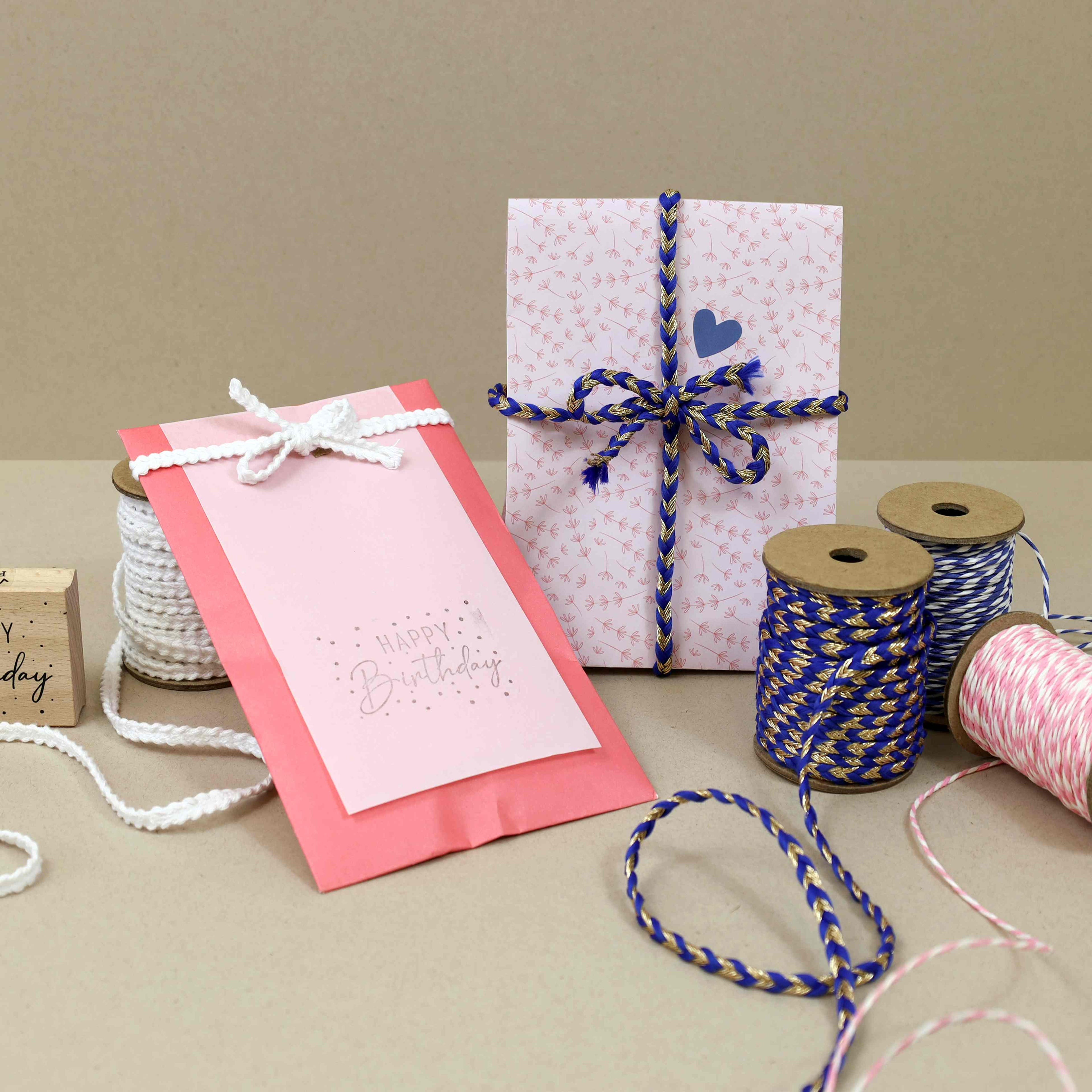 Zwei Geschenkpakete, eines in Rosa und eines mit einem Herz, sind mit geflochtener Schnur verziert. Auf einer beigen Fläche befinden sich Spulen mit buntem Garn und eine "Happy Birthday"-Karte.