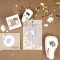 Matériel de bricolage d'Artoz Papier AG sur une surface brune, idéal pour les Basteln : comprend le Stanzer S Glückschwein, la perforatrice licorne, une enveloppe décorée, une carte de vœux, un paquet cadeau, des confettis, des tiges de fleurs et des découpes de Glückschwein.