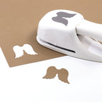 Eine weiße Artoz Papier AG Stanzer M Engelsflügel Bastelstanze stanzt Flügelformen aus braunem Papier für Basteln. Ein brauner Flügelausschnitt liegt auf einer weissen Fläche, ein anderer bleibt im Blatt - perfekt zum Kartenbasteln.
