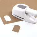 Une étiquette Artoz Papier AG Stanzer M Hangtag est découpée en forme d'étiquette avec des bords festonnés dans une feuille brune pour Karten Basteln. Une étiquette finie se trouve à proximité sur la surface blanche, mettant en valeur son design distinct.