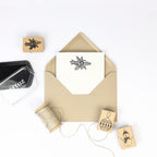 Ein brauner Umschlag mit einer Blumenkarte ist umgeben von Artoz Papier AG Bastelarbeiten: einer Garnrolle, Stempel Gesteck Holzstempel (Tannenzweig, Ornament, Schneemann) und einem Stempelkissen - perfekt für einzigartige Einladungskarten.
