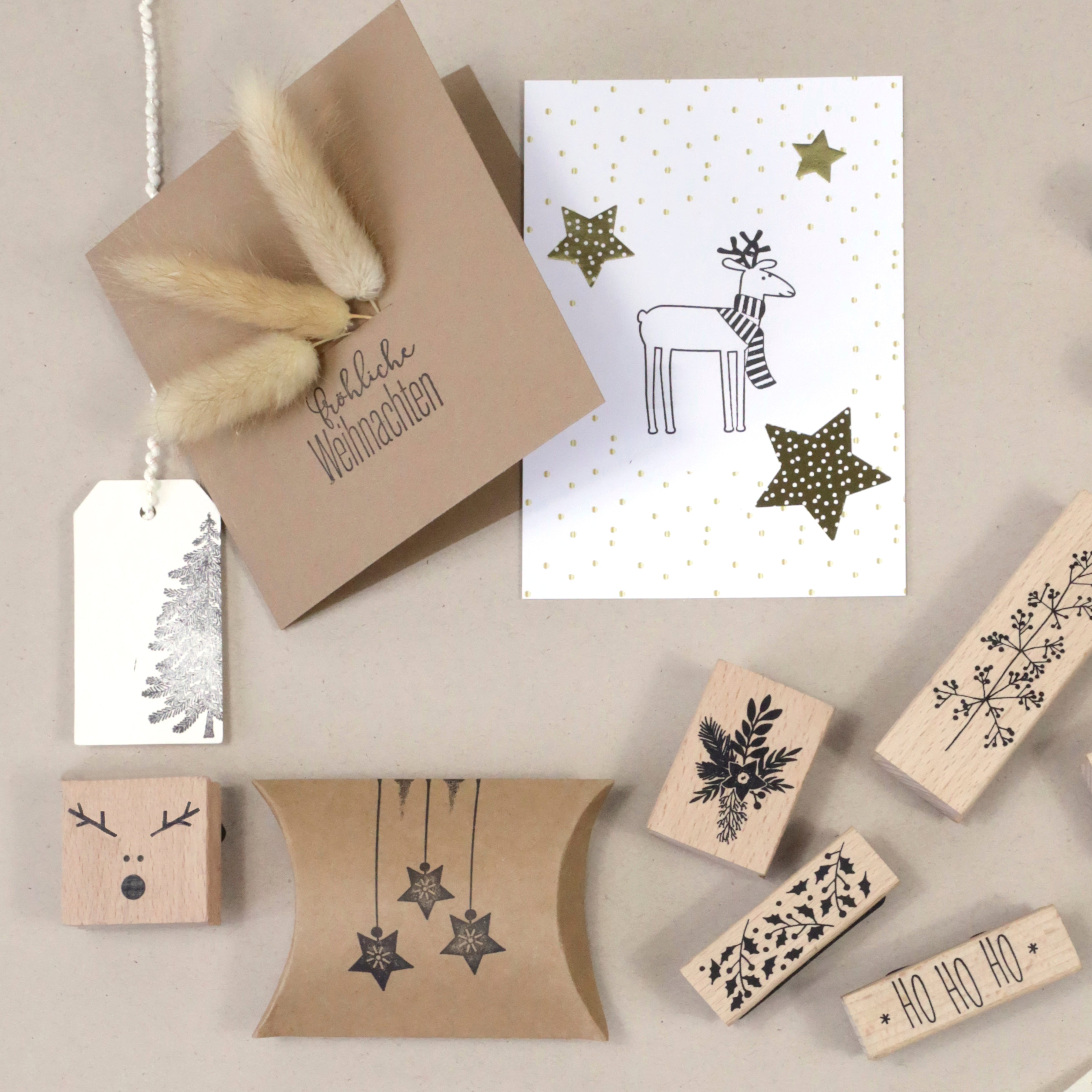 Ein Flat Lay von weihnachtlichen Bastelarbeiten mit dem Stempel Sterne hängend der Artoz Papier AG, festlichen Geschenkanhängern, einer Grußkarte mit Sternemotiv und Rentieren, einer Kissenschachtel und Holzstempeln auf hellbeigem Hintergrund.