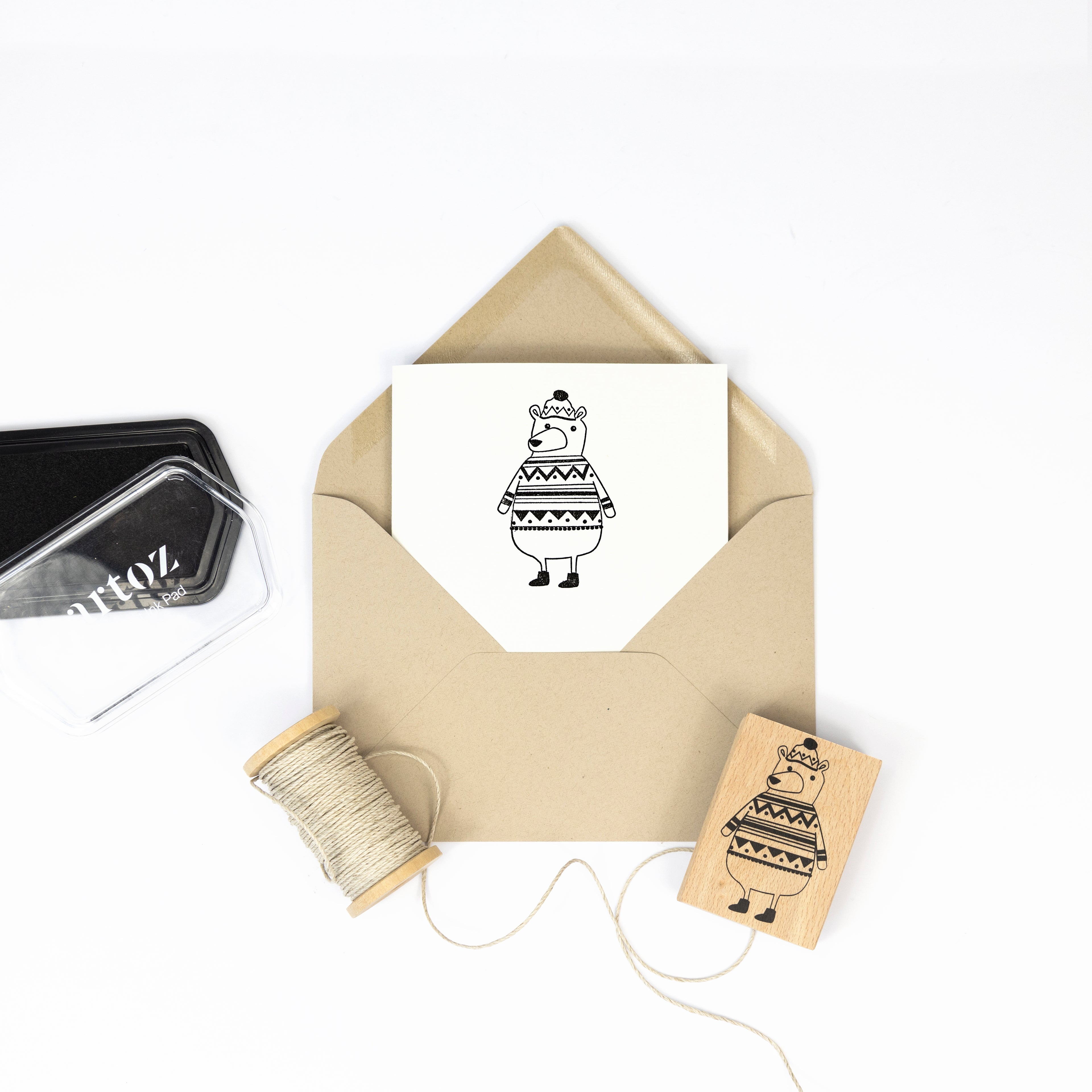 Ein brauner Umschlag mit einer Karte im Inneren, auf der eine schwarze Strichzeichnung eines Bären in einem Pullover abgebildet ist. In der Nähe: Bastelarbeiten-Zubehör-Artoz Papier AG Stempel Winterbär, passendes Motiv, Schnurspule und schwarzes Stempelkissen.