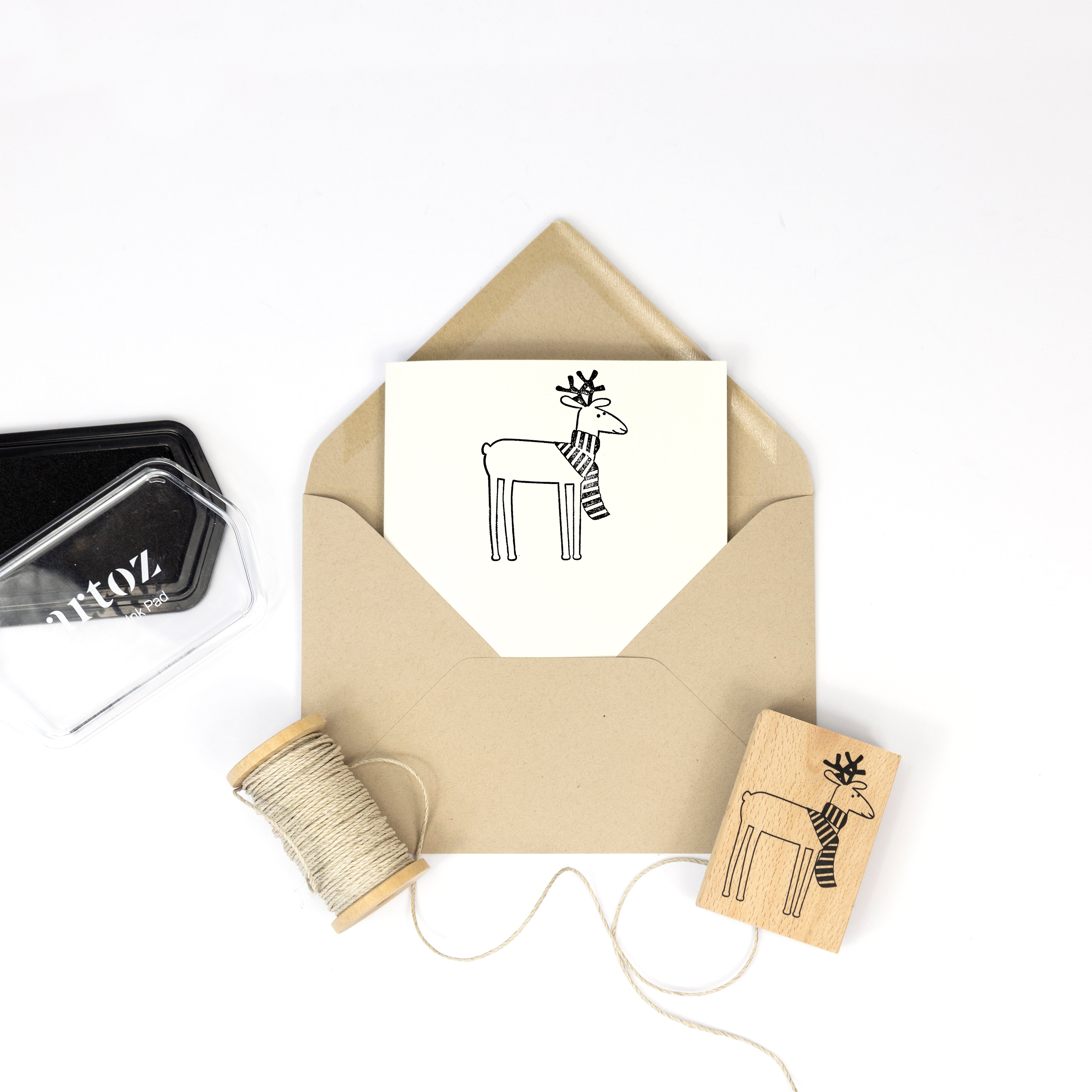 Une enveloppe brune contient une carte représentant un renne avec une écharpe. À proximité se trouvent les éléments de base du Bastelarbeiten : ficelle, tampon encreur noir et le Stempel Renntier d'Artoz Papier AG, qui reprend le même adorable motif.