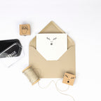 Ein brauner Umschlag mit einer Karte, die ein minimalistisches Rentiergesicht zeigt, sowie Bastelarbeiten-Materialien: Bindfaden, Einladungskarten und der Artoz Papier AG Stempel Rentierkopf - alles auf weißem Hintergrund.