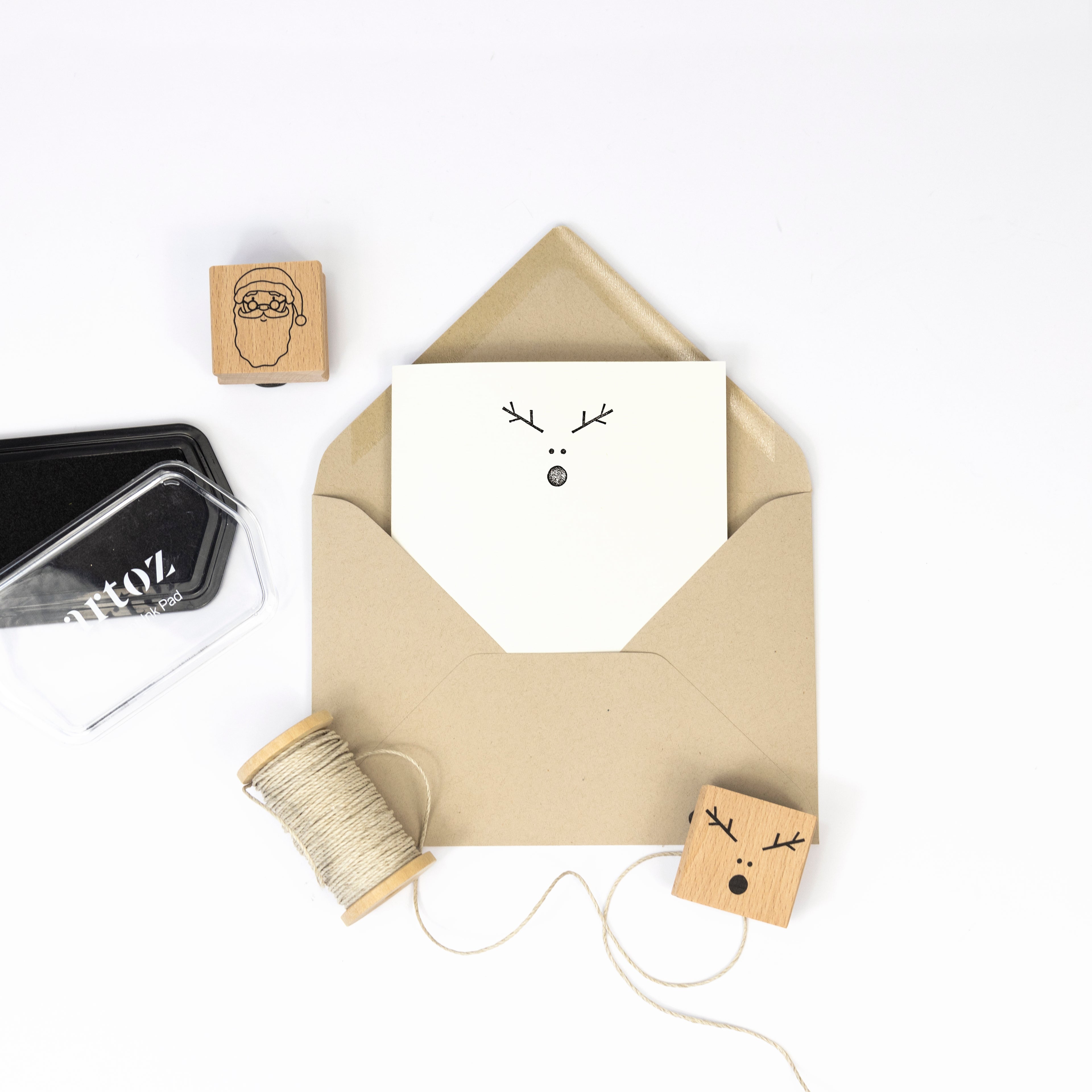 Ein brauner Umschlag mit einer Karte, die ein minimalistisches Rentiergesicht zeigt, sowie Bastelarbeiten-Materialien: Bindfaden, Einladungskarten und der Artoz Papier AG Stempel Rentierkopf - alles auf weißem Hintergrund.