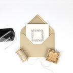 Une Einladungskarten carrée avec une bordure florale est placée dans une enveloppe marron. À proximité se trouvent un tampon encreur noir, de la ficelle Bastelarbeiten et le Stempel Bordüre Eckig d'Artoz Papier AG, tous ornés de motifs assortis sur fond blanc.