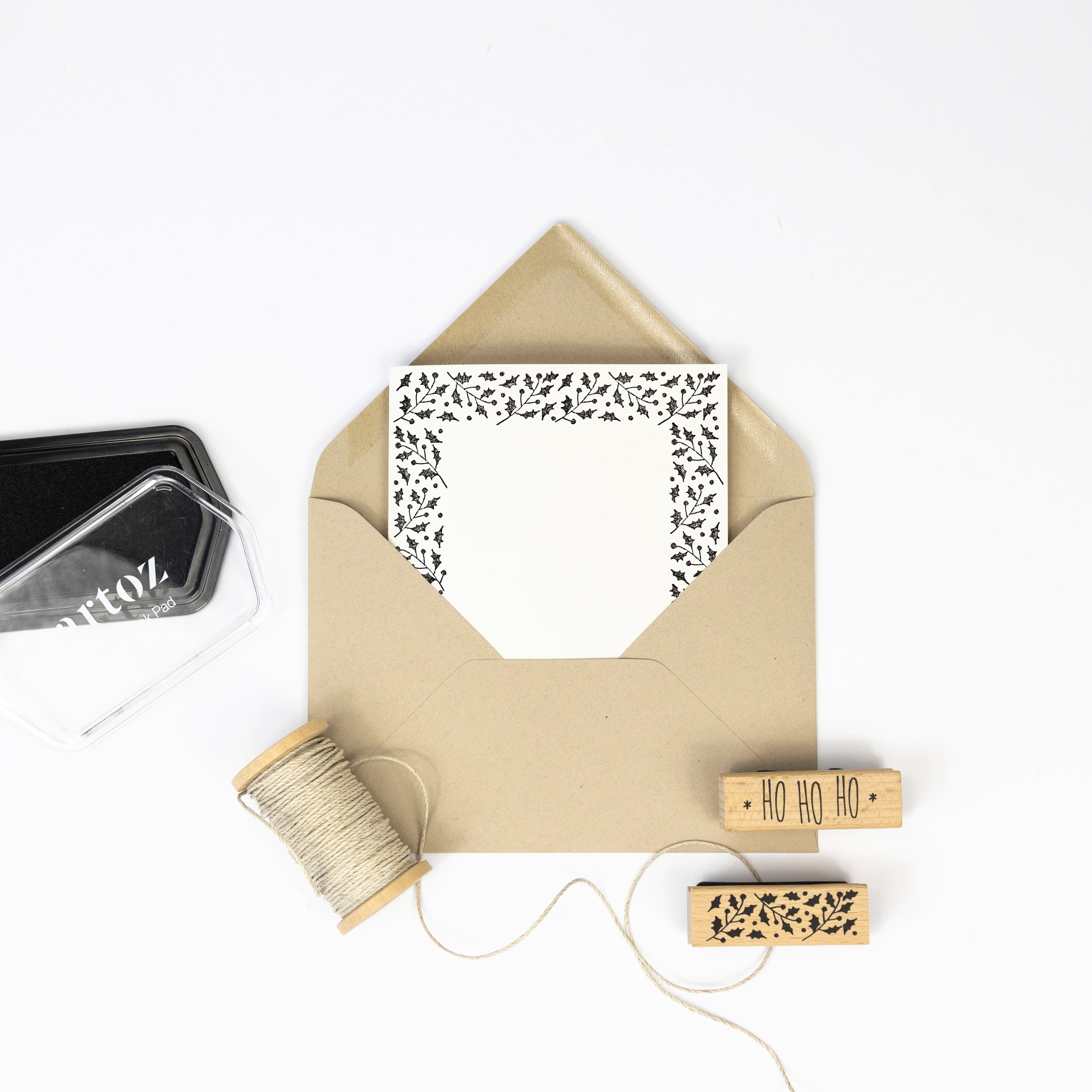 Une enveloppe en papier kraft contenant une carte décorative est entourée des éléments essentiels de Bastelarbeiten : de la ficelle, un tampon encreur noir et deux Artoz Papier AG Stempel Stechpalme, dont l'un est estampillé "HO HO HO", le tout présenté sur un fond blanc.