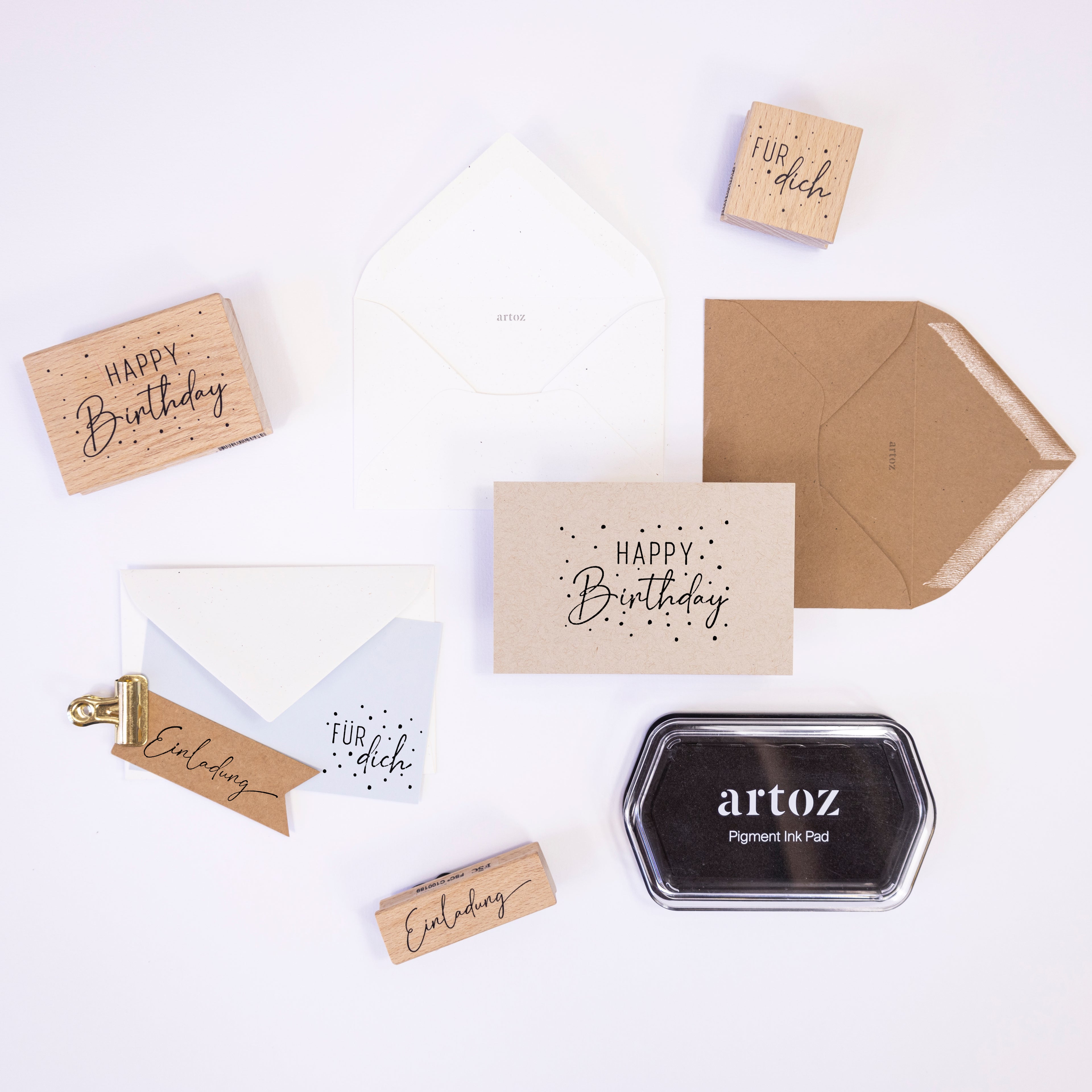 Une présentation plate comprenant des cartes Artoz Papier AG Stempel Einladung, des enveloppes, un Holzstempel avec des textes en allemand et en anglais, une pince à reliure dorée et un tampon d'encre pigmentaire noir Artoz - idéal pour les travaux manuels créatifs.