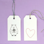 Zwei weiße Artoz Papier AG Geschenkanhänger auf lila: ein Anhänger mit dem Stempel Glücksschweinchen mit Blumen, der andere mit einem umrandeten Herz. Beide haben Schnurschlaufen - ideal zum Basteln oder Verschenken.