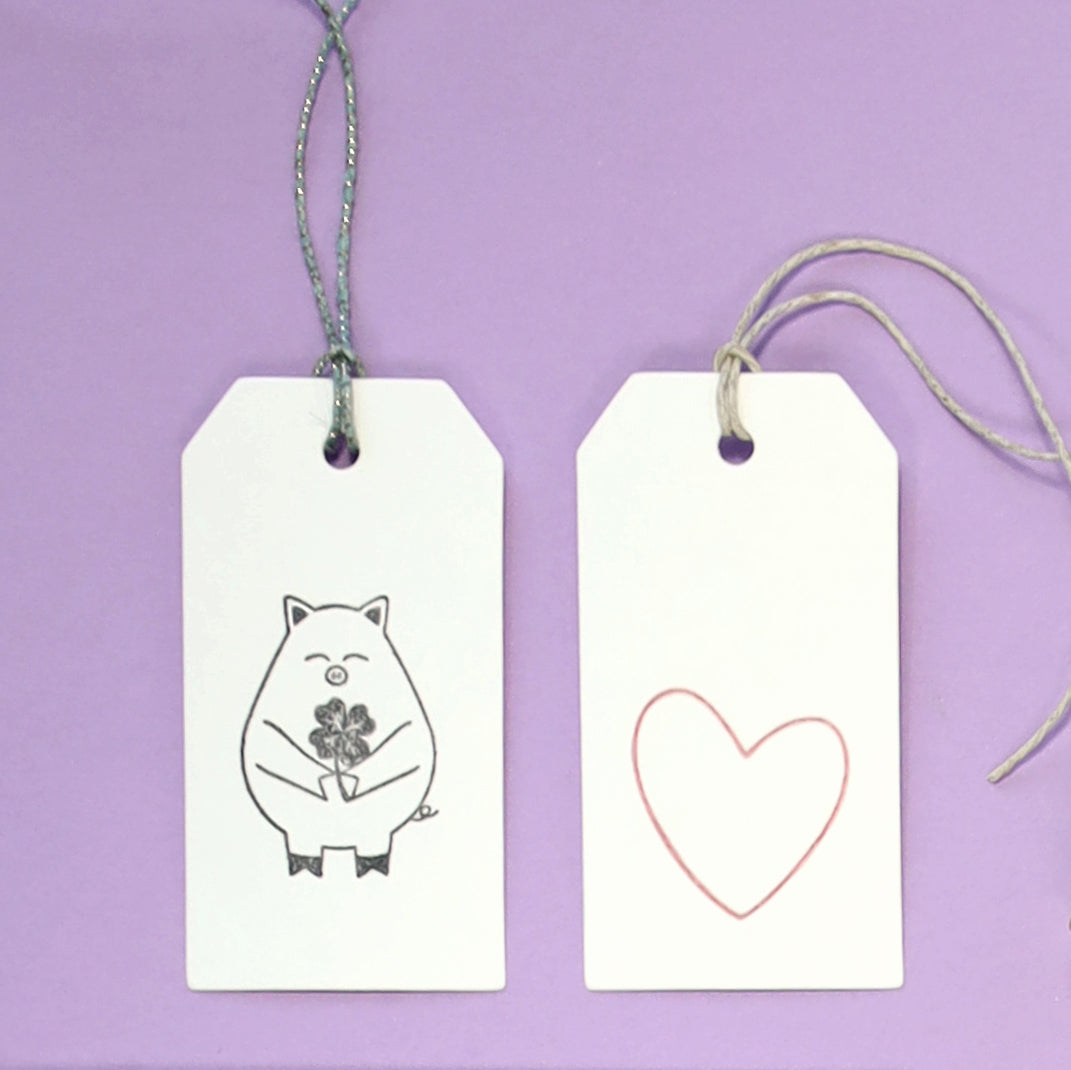 Zwei weiße Artoz Papier AG Geschenkanhänger auf lila: ein Anhänger mit dem Stempel Glücksschweinchen mit Blumen, der andere mit einem umrandeten Herz. Beide haben Schnurschlaufen - ideal zum Basteln oder Verschenken.