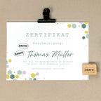 Ein deutsches Zertifikat auf einem Klemmbrett mit sechseckigen Motiven, mehrfach gestempelt mit dem Artoz Papier AG Stempel Bravo! und dem Namen Thomas Müller in grosser Schrift, das hervorragende Leistungen bei Bastelarbeiten und die Erfüllung aller Anforderungen bescheinigt.