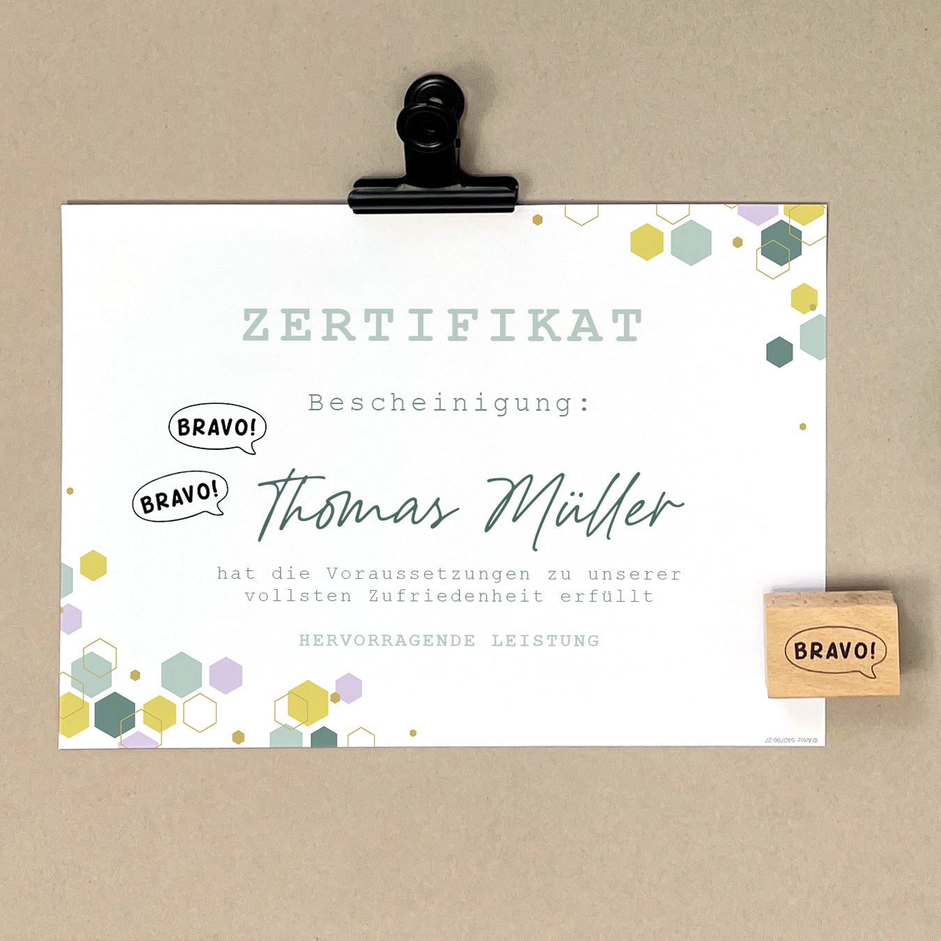 Ein deutsches Zertifikat auf einem Klemmbrett mit sechseckigen Motiven, mehrfach gestempelt mit dem Artoz Papier AG Stempel Bravo! und dem Namen Thomas Müller in grosser Schrift, das hervorragende Leistungen bei Bastelarbeiten und die Erfüllung aller Anforderungen bescheinigt.