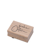 Der Artoz Papier AG Stempel Frohe Ostern ist ein rechteckiger Holzstempel für Bastelarbeiten, auf dem handschriftlich "Frohe Ostern" und ein einfaches Osterei mit Gesicht auf weißem Hintergrund abgebildet sind.