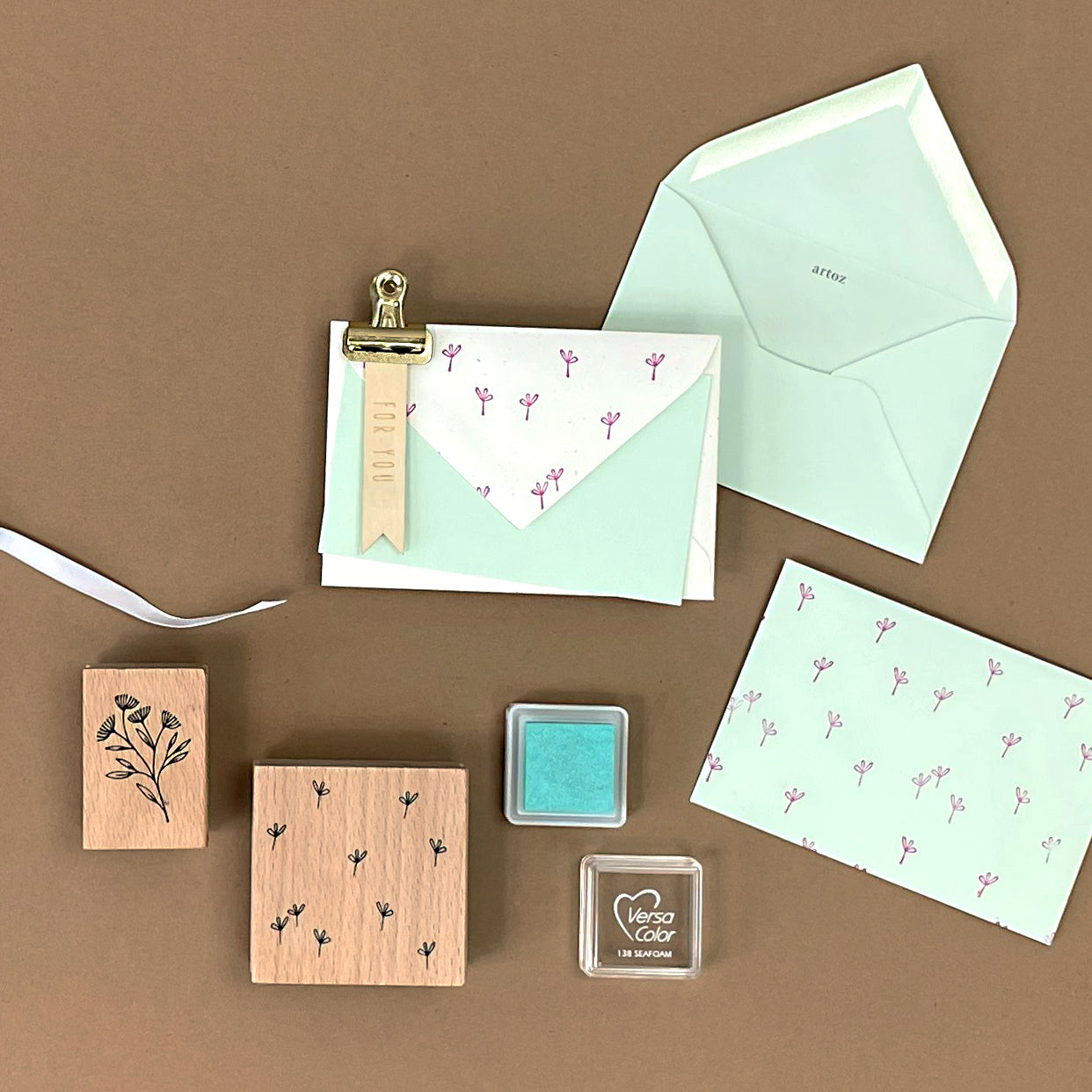 Ein flaches Layout mit mintgrünen Umschlägen, rosa Blumen-Notizkarten, Bastelarbeiten wie dem Artoz Papier AG Stempel Blumen all over Holzstempel, einem blaugrünen Stempelkissen, einer Metallklammer, einem Band und einem gestempelten Holzanhänger auf einer braunen Oberfläche.