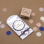 Ein dekorativer Umschlag und ein Artoz Papier AG Stempel Kreis mit Text, beschriftet für die Freudentränen, liegen auf einer braunen Fläche mit blauem, weißem und rosa Konfetti - ideal für kreative Bastelarbeiten.