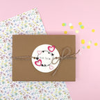 Une boîte cadeau marron avec un nœud en ficelle et une étiquette ronde "Pour toi", décorée de cœurs roses estampillés à l'aide du Stempel Kreis mit Herz d'Artoz Papier AG, repose sur du papier fleuri et des confettis roses - idéal pour les Bastelarbeiten.