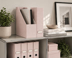 Une étagère soigneusement agencée, sur laquelle sont disposés des porte-revues Artoz Papier AG 2er Stehsammler roses, des boîtes, des classeurs, une plante en pot verte, des livres empilés, un bol et une œuvre d'art abstrait encadrée, crée une ambiance minimaliste et élégante de bureau à domicile.