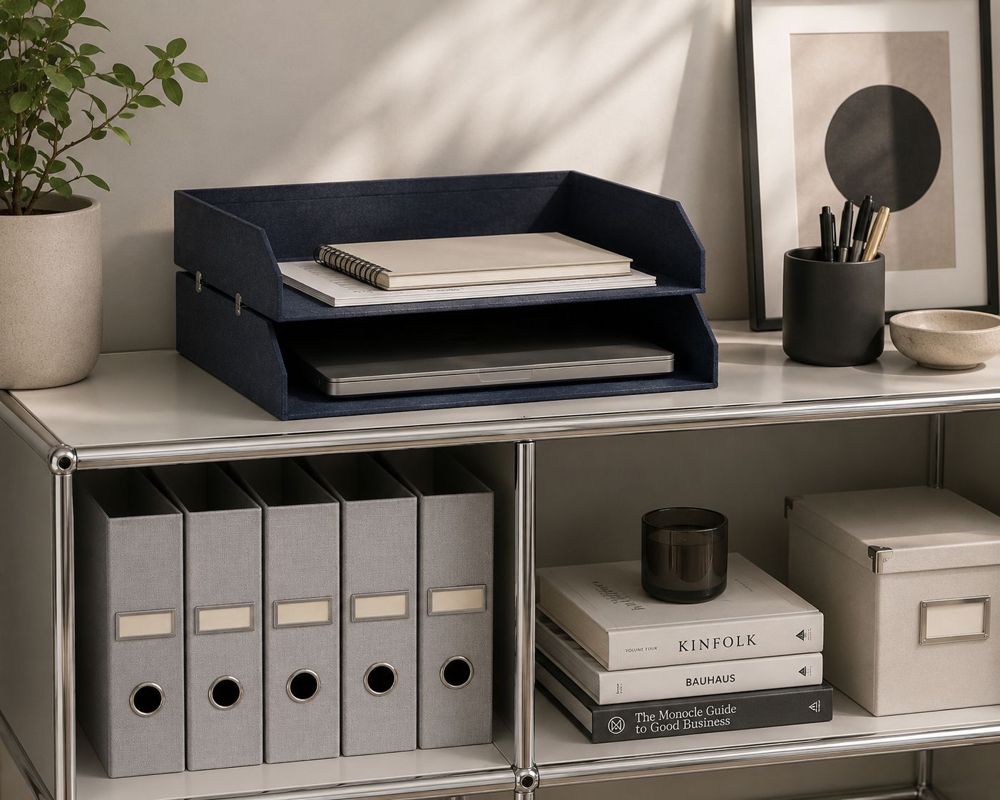 Une étagère de bureau bien rangée, équipée de boîtes à dossiers grises et du plateau « Ablagefach doppelt » d'Artoz Papier AG, accueille un ordinateur portable, des livres, une estampe abstraite, une bougie, une plante et un pot à stylos : l'équipement idéal pour un bureau à domicile soigné et minimaliste.