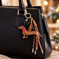 Un sac à main noir chic doté de fermetures éclair dorées est orné du « Anhänger Hund aus PU - Lederoptik » d'Artoz Papier AG, composé d'un ravissant pendentif en forme de teckel marron, d'un cœur noir et de pompons marrons suspendus à l'une des anses. Un éclairage tamisé et un arrière-plan flou renforcent l'ambiance chaleureuse, faisant de cet accessoire une adorable idée de cadeau pour les amateurs de chiens.