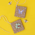 Zwei braune quadratische Anhänger mit weißen Herzchen, ein lila Artoz 3D Sticker Schmetterling der Artoz Papier AG und eine grüne Blume sind Schweizer Handarbeit mit Schnurschlaufen. Zwei kleine Bienenaufkleber sitzen auf einem leuchtend gelben Hintergrund.