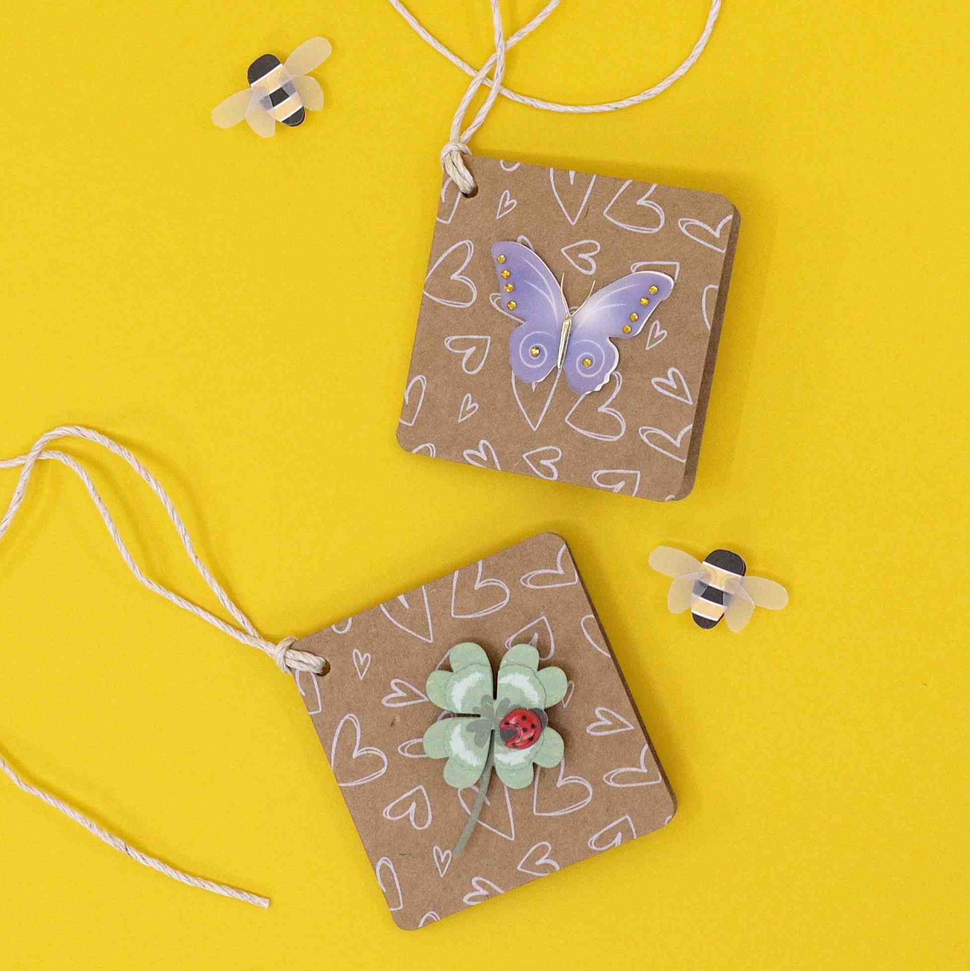 Deux étiquettes carrées brunes avec des gribouillis en forme de cœur blanc, un autocollant violet Artoz 3D Sticker Schmetterling d'Artoz Papier AG et une fleur verte sont des créations suisses faites à la main avec des boucles de ficelle. Deux petites abeilles autocollantes se trouvent sur un fond jaune vif.