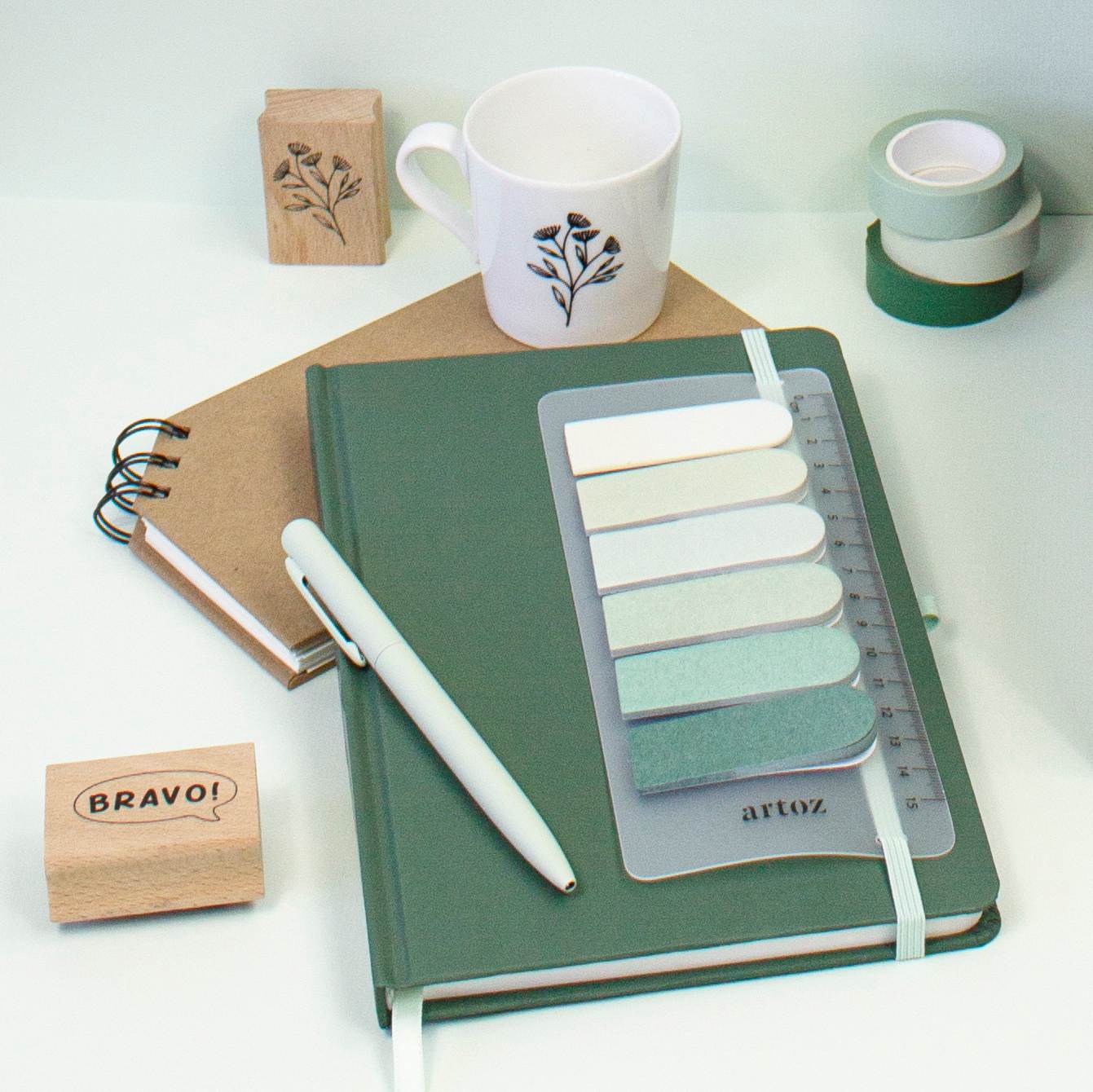 Das Artoz Papier AG Notizbuch A5 mit Kugelschreiber, Haftnotizen, ein Spiralnotizbuch, eine Blumentasse, zwei Stempel und drei Rollen Washi-Tape in Grün- und Weißtönen.