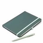 Das Notizbuch A5 mit Kugelschreiber von Artoz Papier AG in Dunkelgrün mit Punktmuster und hellblauem Gummiband liegt auf weißer Fläche neben dem passenden Stift.