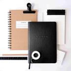 Un ensemble d'articles de papeterie : un carnet de notes A5 à spirale marron avec couverture rigide, un carnet de notes A5 Artoz Papier AG noir, une règle, un stylo, un rouleau de ruban adhésif blanc, un bloc-notes avec du papier ligné et des cartes de notes, le tout sur une surface blanche.