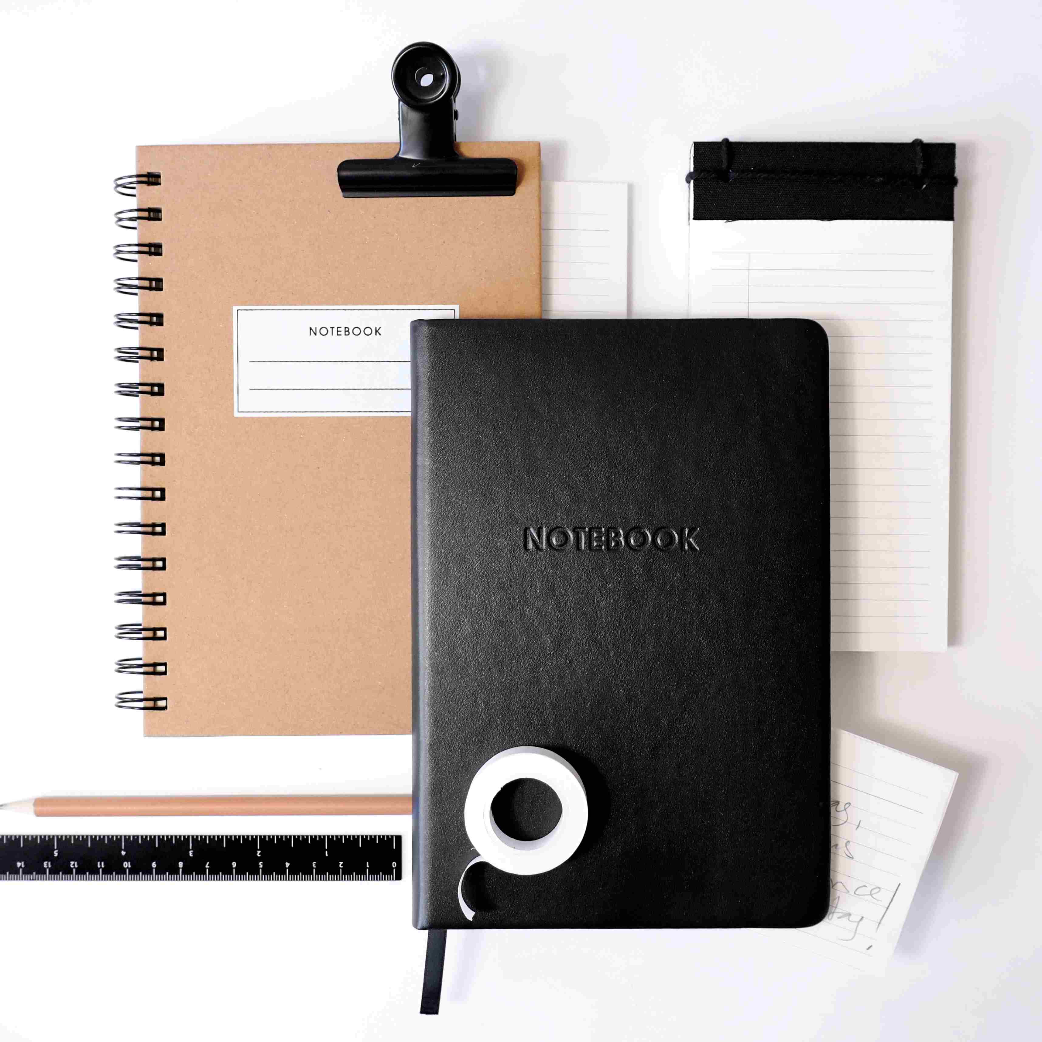 Un ensemble d'articles de papeterie : un carnet de notes A5 à spirale marron avec couverture rigide, un carnet de notes A5 Artoz Papier AG noir, une règle, un stylo, un rouleau de ruban adhésif blanc, un bloc-notes avec du papier ligné et des cartes de notes, le tout sur une surface blanche.