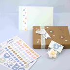 Ein pastellfarbenes Geschenkset mit einer verzierten Karte, einer braunen Geschenkschachtel mit Schleife und "For You"-Anhänger, Artoz Sticker Gans von Artoz Papier AG zur Dekoration und weißen Bonbons, die alle auf einer hellblauen Fläche angeordnet sind.