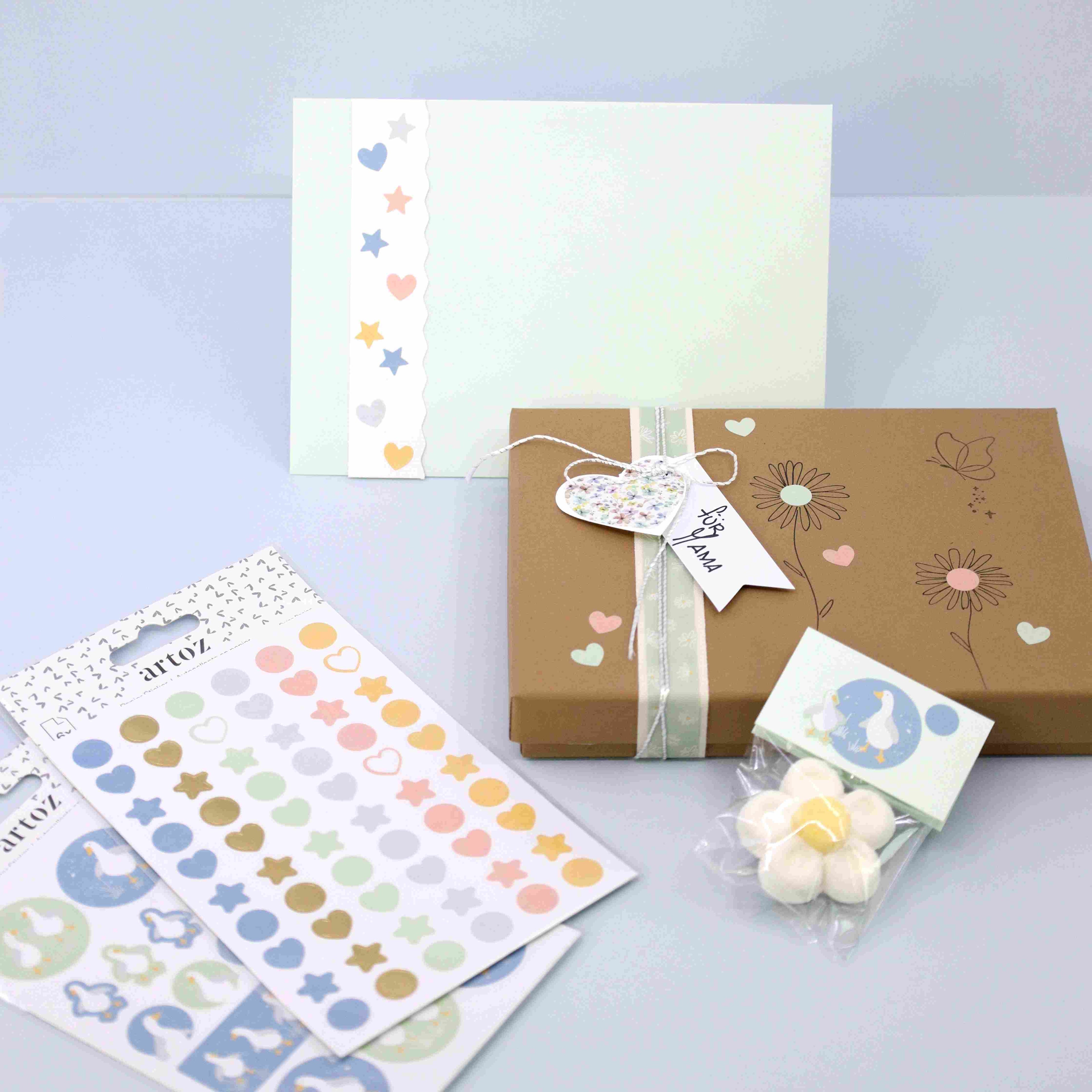 Un coffret cadeau au thème pastel comprenant une carte décorée, une boîte cadeau marron avec ruban et étiquette "For You", Artoz Sticker Gans by Artoz Papier AG pour la décoration, et des bonbons blancs, le tout disposé sur une surface bleu clair.