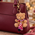 Ein Teddy-Anhänger – ein Plüschbär mit Halstuch von Artoz Papier AG – baumelt am Henkel einer bordeauxroten Ledertasche, die auf einem Tisch steht, und macht sie zu einer charmanten Weihnachtsgeschenkidee.