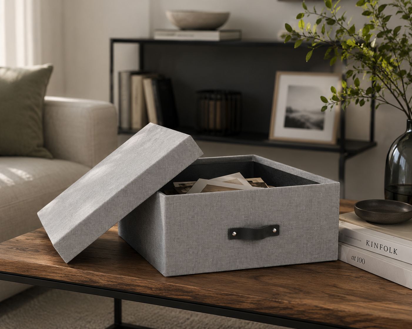 Une boîte de rangement ouverte et grise de la marque Artoz Papier AG, à l'aspect lin et dotée d'une poignée noire, repose sur une table basse en bois et contient des photos et des papiers. À l'arrière-plan, on aperçoit un canapé, une étagère décorative et une plante verte : l'ensemble est idéal pour créer un espace de rangement élégant.