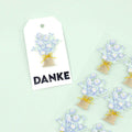 Une étiquette cadeau blanche avec un bouquet floral bleu et blanc, une ficelle jaune et "DANKE", présentée à côté des motifs de l'autocollant 3D "Blumenstrauss" d'Artoz Papier AG sur un fond vert clair.
