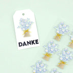 Une étiquette cadeau blanche avec un bouquet floral bleu et blanc, une ficelle jaune et "DANKE", présentée à côté des motifs de l'autocollant 3D "Blumenstrauss" d'Artoz Papier AG sur un fond vert clair.