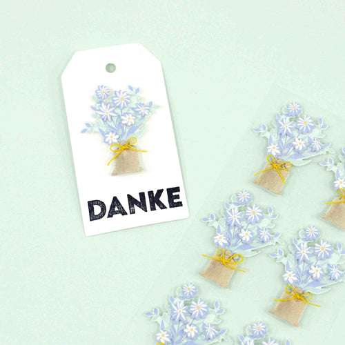 Une étiquette cadeau blanche avec un bouquet floral bleu et blanc, une ficelle jaune et "DANKE", présentée à côté des motifs de l'autocollant 3D "Blumenstrauss" d'Artoz Papier AG sur un fond vert clair.