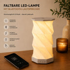 Die faltbare LED-Tischlampe „Sona“ von Artoz Papier AG mit dimmbarem warmweißem Licht, spiralförmigem Lampenschirm und integriertem Bluetooth-Lautsprecher steht auf einem Holztisch neben einem Smartphone, einer Kerze und einer Pflanze. Symbole heben die Funktionen hervor.