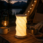 Eine leuchtende faltbare LED-Tischlampe „Sona“ mit Bluetooth-Lautsprecher von Artoz Papier AG steht auf einem Holztisch neben Bechern, Nüssen und einer Vintage-Laterne; dahinter sind ein Zelt, eine Lichterkette und ein See vor einer Bergkulisse in der Dämmerung zu sehen.