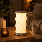 Eine faltbare LED-Tischlampe „Sona“ mit Bluetooth-Lautsprecher von Artoz Papier AG steht auf einem Holztisch neben einer Kerze, einer Tasse und einem Buch. Im Hintergrund sorgt eine Decke über dem Sofa für eine gemütliche Atmosphäre.