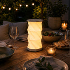 Sur une table en bois installée en plein air se trouve la lampe de table LED Aurora d'Artoz Papier AG, réglable en intensité et pliable, dotée d'une commande tactile et d'un port USB-C, entourée de vaisselle, d'une bougie, d'un saladier et de guirlandes lumineuses, au milieu de la verdure.