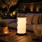 Die faltbare LED-Laterne „Timber“ aus Holz von Artoz Papier AG spendet dimmbares, warmweißes Licht auf einem Holztisch voller Getränke und Snacks. Im Hintergrund sorgen Sofa und beleuchtete Pflanzen für eine gemütliche Atmosphäre.