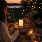 Une femme reçoit la lampe de table LED pliable Minora d'Artoz Papier AG — à intensité variable, avec commande tactile et port USB-C — lors d'un pique-nique au coucher du soleil, entourée de mets, de fleurs et de guirlandes lumineuses qui rehaussent le cadre enchanteur.