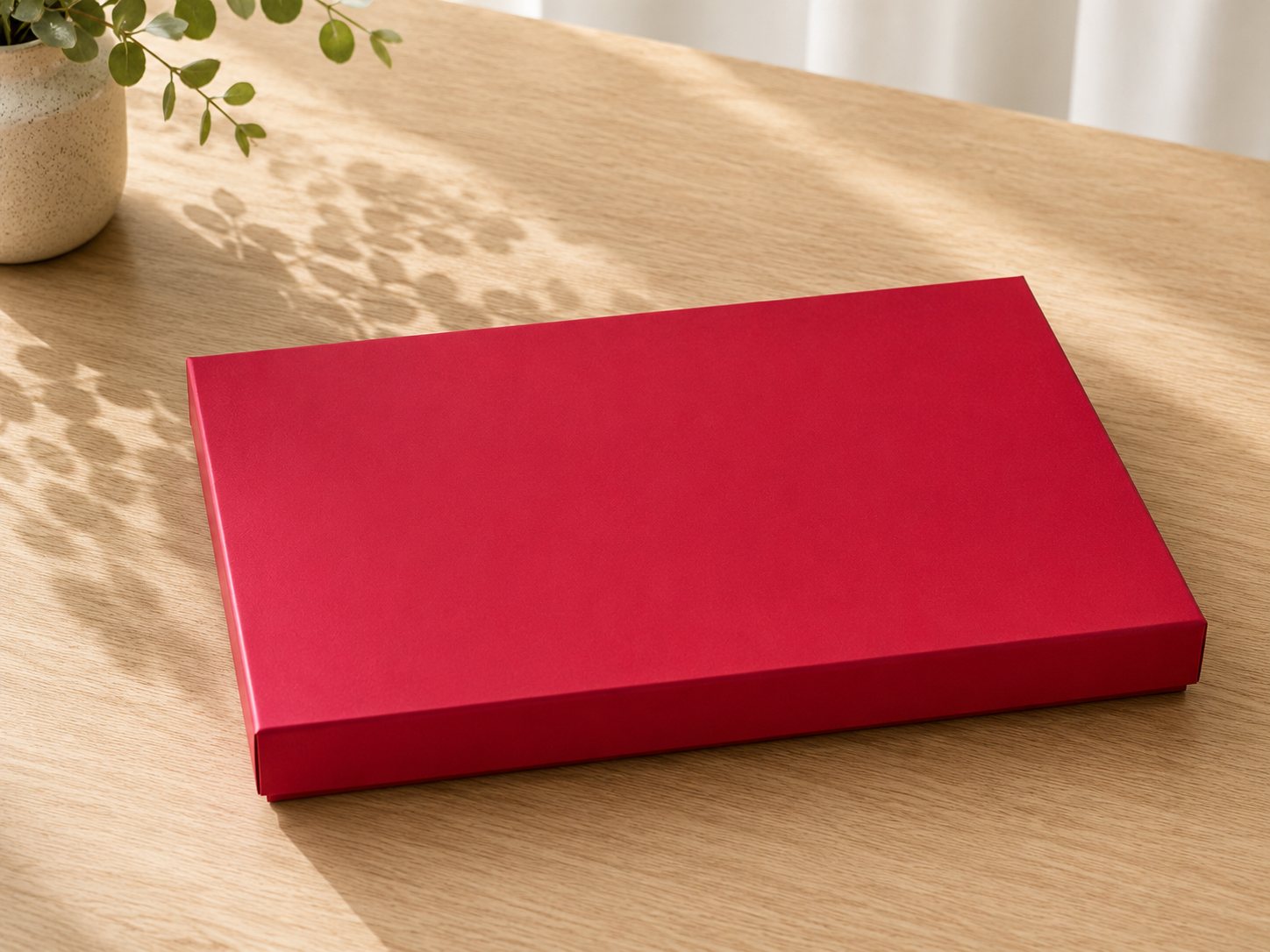 Eine rechteckige, flache, rote Geschenkbox „Pure“ von Artoz Papier AG im A4-Format steht auf einem hellen Holztisch, der von sanftem Tageslicht beleuchtet wird, während im Hintergrund die Schatten von Blattpflanzen zu sehen sind.
