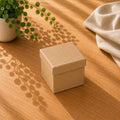 Auf einem Holztisch neben einer Topfpflanze steht eine mittelgroße, geschlossene Geschenkbox „Pure“ von Artoz Papier AG; Sonnenlicht wirft sanfte Blattschatten, und im Hintergrund ist ein leichter Stoff zu sehen.