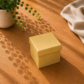Une boîte cadeau « Geschenk-Box Pure » de taille moyenne, carrée et jaune, de la marque Artoz Papier AG, repose fermée sur une table en bois à côté d'une plante en pot ; la lumière du soleil projette des ombres de feuilles et un tissu beige est visible en arrière-plan.