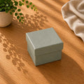 Une boîte-cadeau « Pure » taille M de la marque Artoz Papier AG, de couleur grise et fermée, repose sur une table en bois sur laquelle se dessinent les ombres des plantes, accompagnée d'une plante en pot et d'un tissu beige. Une douce lumière du soleil projette des lignes diagonales sur la scène.