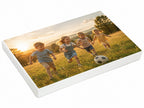Vier kleine Kinder rennen lachend umher, während sie bei Sonnenuntergang auf einer Wiese Fußball spielen – im Hintergrund sind Bäume zu sehen, vor ihnen liegt ein Fußball. Ein fröhlicher Moment, der sich perfekt für die „Personalisierte Geschenkbox A4“ – mit eigenem Bild und/oder Text gestalten von Artoz Papier AG, wodurch diese wertvolle Erinnerung zu einem wahrhaft einzigartigen Geschenk wird.