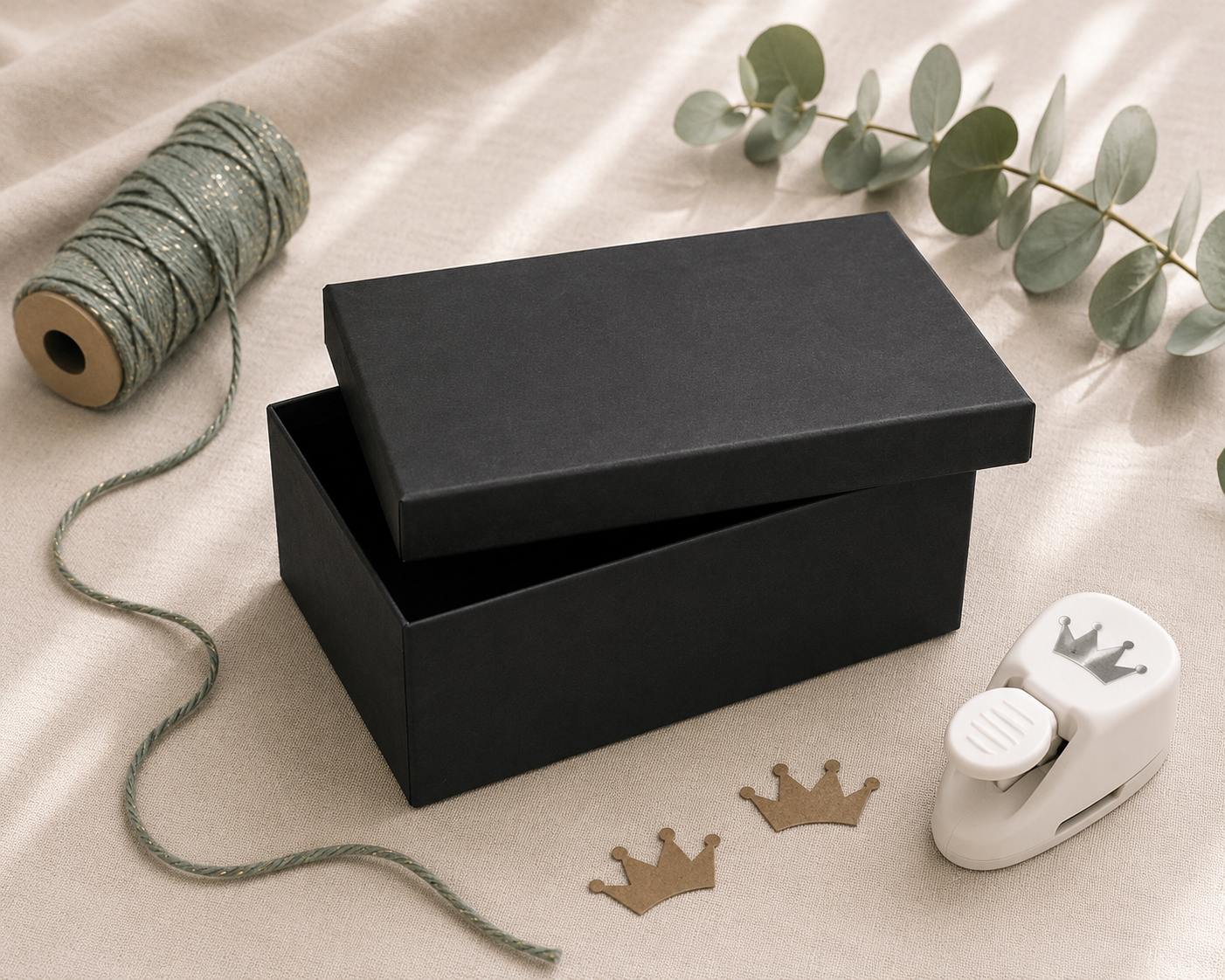 Eine geöffnete schwarze Geschenkbox „Pure L“ (rechteckig) von Artoz Papier AG liegt auf einem beigen Tuch, umgeben von grünem Bindfaden, Eukalyptusblättern, einem kronenförmigen Papierstanzer und zwei aus braunem Papier ausgestanzten Kronen.