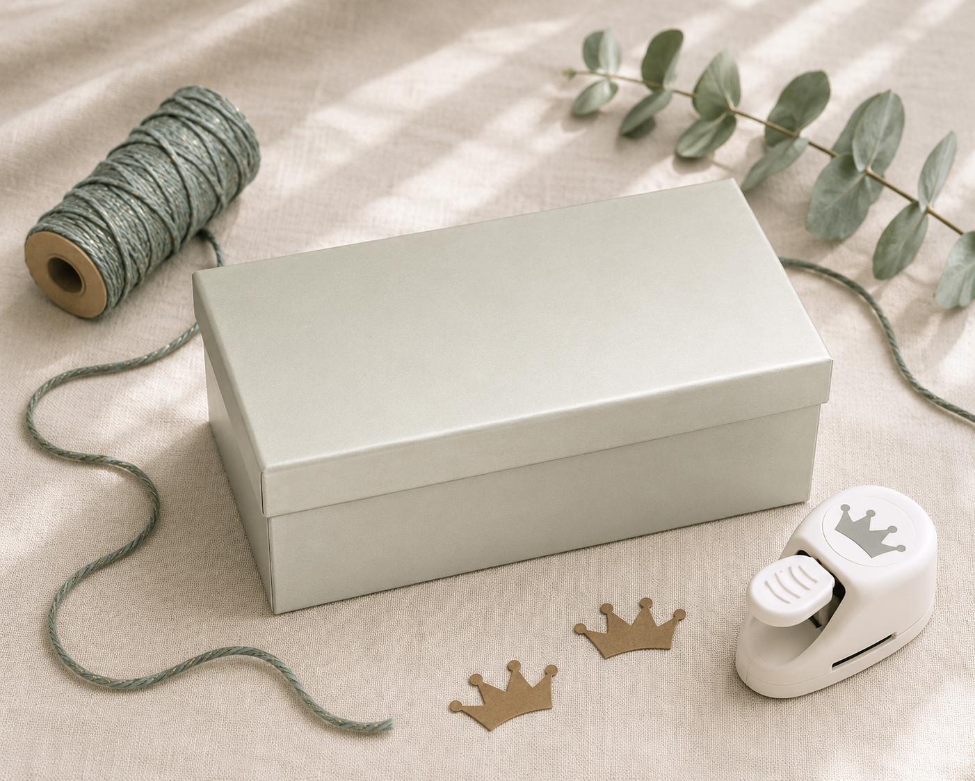 Eine rechteckige Geschenkbox „Pure L“ der Artoz Papier AG in Hellgrau liegt geschlossen auf einem Stoff, umwickelt mit einer grünen Schnur, einer Spule, Eukalyptusblättern, einer weißen Bastelstanze mit Kronenmotiv und zwei Papierkronen – ein bezauberndes Arrangement, das sich ideal zum kreativen Verpacken von Geschenken eignet.
